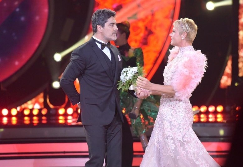 Para entrar no clima do dia dos namorados, Xuxa recebeu Junno no palco com um vestido branco, clássico e rendado. Para complementar o look, ela apostou em mangas cor de rosa, que deram um toque especial