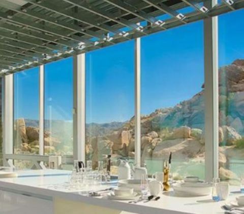 A cozinha tem piso de concreto e superfícies de mármore, com fornos duplos, freezer, geladeira e eletrodomésticos sofisticados. Bancadas com vista da natureza exterior. 