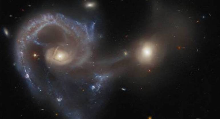Essa fotografia mostra o objeto celeste Arp 107, duas galáxias em processo de fusão. A maior é conhecida como galáxia Seyfert, extremamente energética e com núcleos galácticos ativos. A menor está conectada a ela por uma ponta feita quase que inteiramente de poeira e gás
