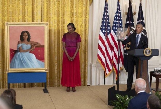 Já na pintura de Michelle Obama, por Sharon Sprung, a ex-primeira-dama posa em um sofá na Sala Vermelha da Casa Branca, usando um vestido de gala azul-claro. Seu olhar é direto e sua expressão tem uma pitada de humor