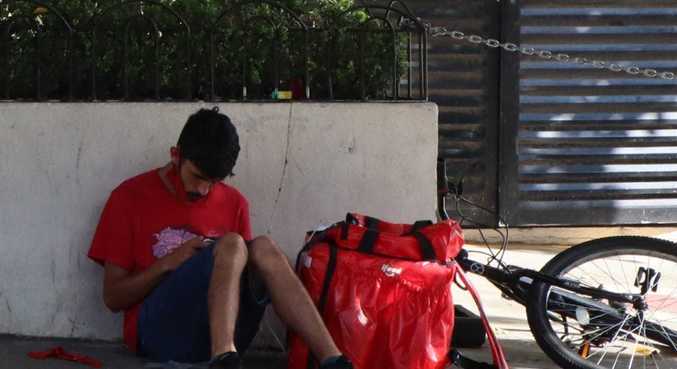 Entregador sentado na calçada de shopping, descansando enquanto espera app solicitar uma entrega