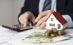 Casa em miniatura, moedas e cédulas de euros sobre uma mesa e pessoa ao fundo mexe em calculadora. Freepik