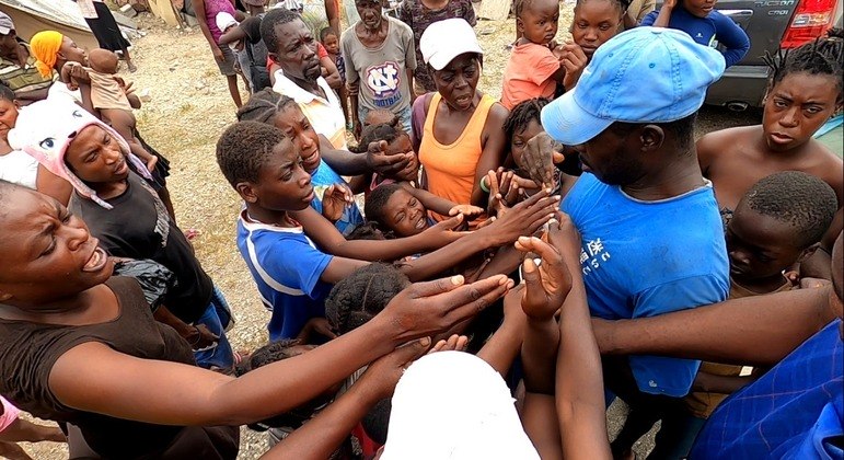 Crianças e mulheres disputam balas distribuídas a partir das mãos do homem com boné azul