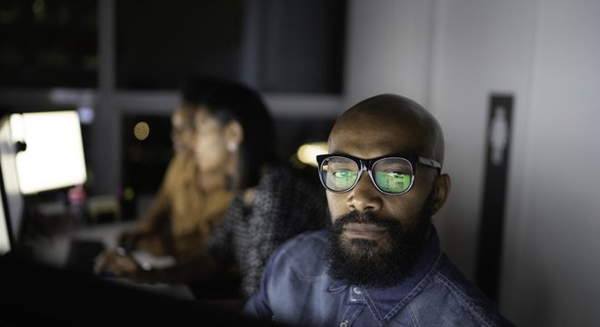 Homem e mulheres no pano de fundo com rostos iluminados por telas de computador em escritório à noite