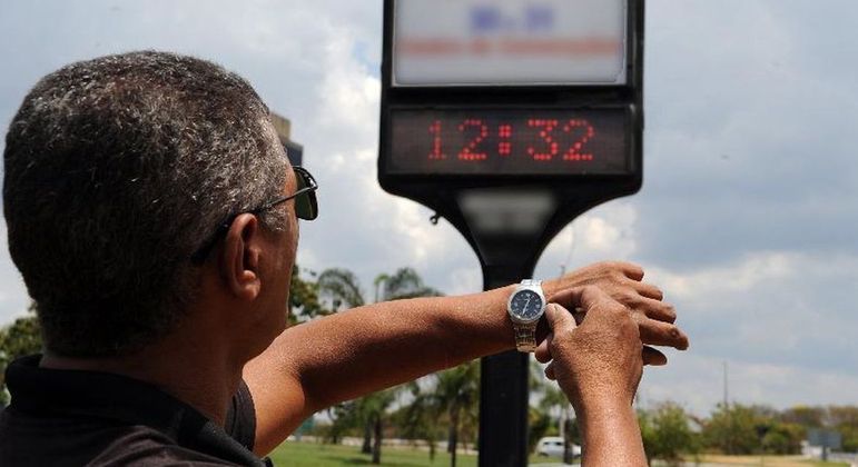 Homem olha relógio de pulso, enquanto relógio de rua marca 12h32; horário de verão pode retornar