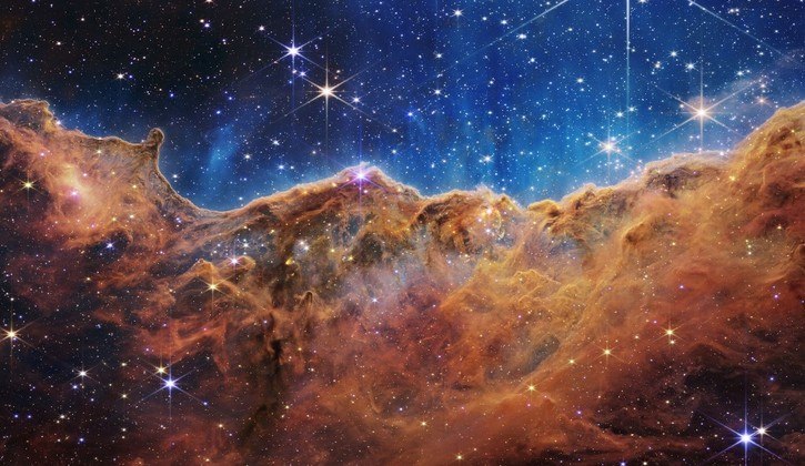 landscape of “mountains” and “valleys” speckled with glittering stars which is actually the edge of a nearby, young, star-forming region called NGC 3324 in the Carina Nebula. Captured in infrared light by the JWST, this image reveals for the first time previously invisible areas of star birth. The JWST is the most powerful telescope launched into space and it reached its final orbit around the sun, approximately 930,000 miles from Earths orbit, in January, 2022. The technological improvements of the JWST and distance from the sun will allow scientists to see much deeper into our universe with greater detail.