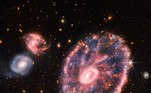 Com a tecnologia infravermelha do telescópio James Webb, a galáxia Cartwheel também foi examinada. Ao lado da grande galáxia rosa, duas galáxias espirais menores aparecem à esquerda. O registro em questão mostra a Cartwheel em estágio transitório; segundo a Nasa, ela ganhou esse formato depois que duas galáxias menores colidiram, transformando-se em uma única