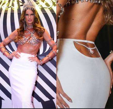 A modelo Izabel Goulart, coroada rainha do Baile do Copa, usou uma longa saia branca de cintura alta e com recorte na parte de trás durante o evento. Um pingente brilhante pendia sobre a região. Ela combinou a peça com um top prateado e brilhoso, que estava em sintonia com a coroa, os braceletes e as pulseiras que compõem o visual 