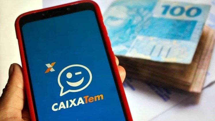 Mão segura celular com o aplicativo Caixa Tem, rodeado por cédulas de reais. Agência Estado / Adriana Toffetti / A7 Press