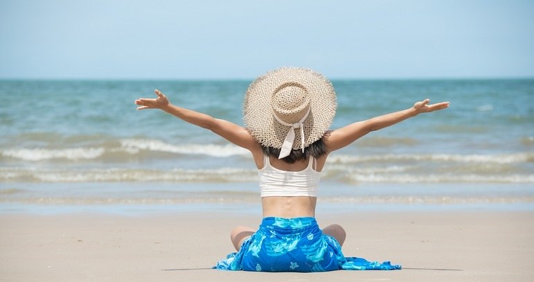 Mulher na praia com chapéu e de braços abertos. Freepik / jcomp