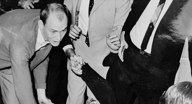 Oswald é levado às pressas para uma ambulância após ser baleado por Jack Ruby