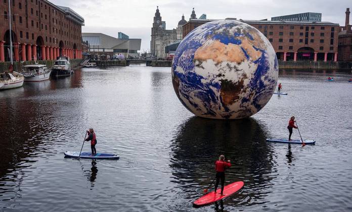 O mundo gira, e o planeta continua maluco! Enquanto isso, pessoas praticam paddle board em torno da instalação de arte 