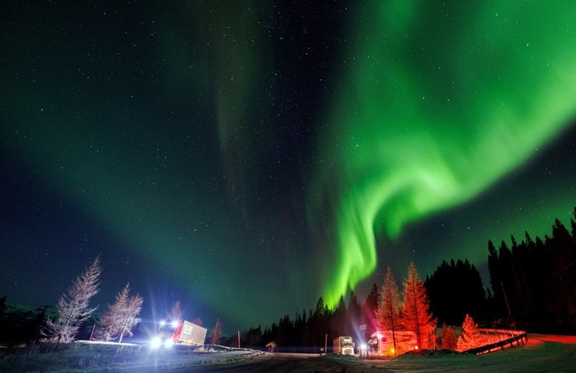 A lindíssima aurora boreal ilumina o céu noturno enquanto pessoas param próximas de rodovias em uma vila da Noruega