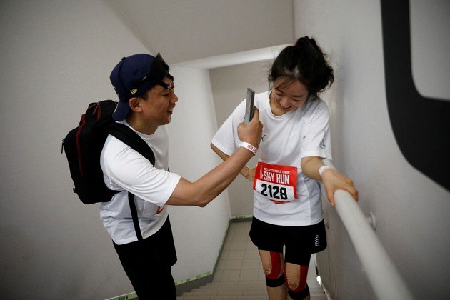 O que está acontecendo aqui?, você deve estar se perguntando. O que parece alguém seguindo algum ídolo, na verdade mostra dois participantes e uma maratona vertical em Seul, na Coreia do Sul, brincando de tirar selfie. Na competição, os maratonistas devem subir 123 andares da Lotte World Tower