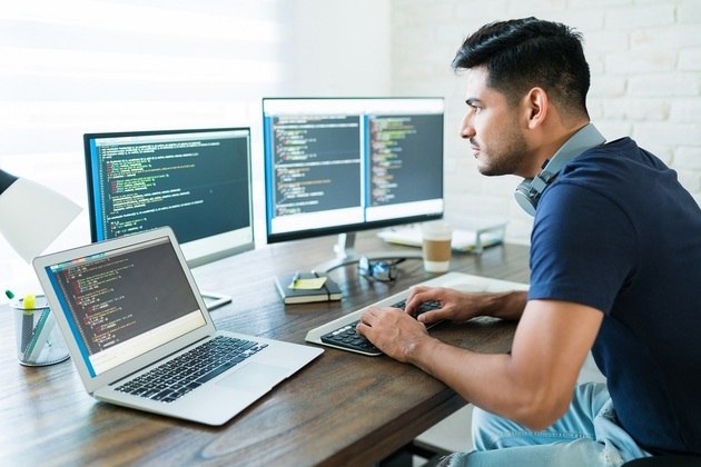 Programador  trabalha em frente a um notebook e dois monitores de computador. Tonodiaz / Freepik