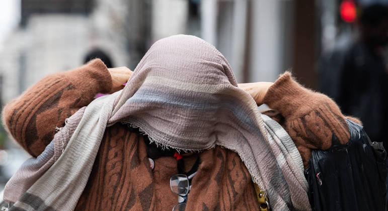 Senhora tenta se proteger do frio ao colocar um lenço no pescoço, mas uma rajada de vento atrapalha
