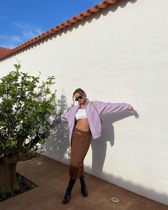 É mais ousadia que você quer? Então, fique com esse look composto de saia longa com fenda na parte de trás, top underboob — que deixa a parte de baixo dos seios à mostra —, jaqueta lilás e botas pretas