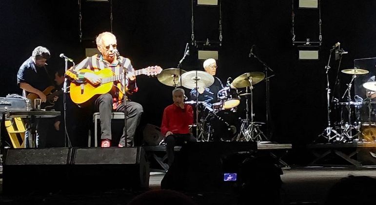 Jards Macalé com seu violão no palco e Caetano sentado ao fundo