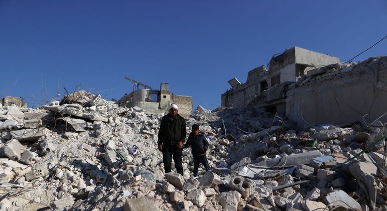 Homem anda com filho sobre escombros de prédio na Síria nesta terça (14)
