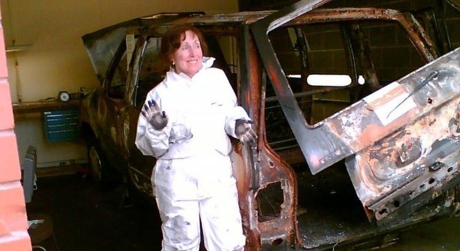 Wiltshire com jaleco, sorrindo e mostrando a mão suja de carvão ao lado de um carro queimado